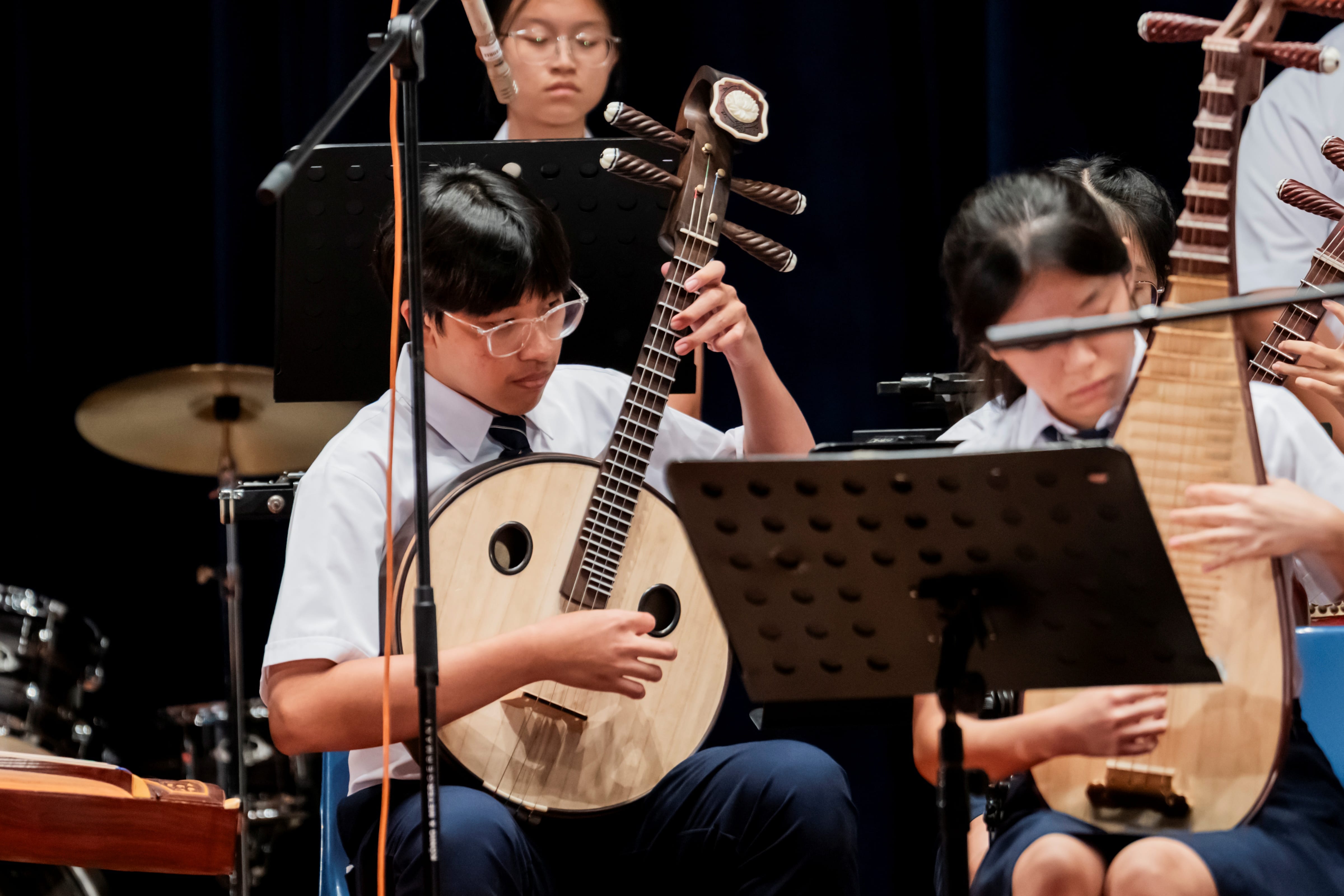 Students in school uniform performing traditional Chinese instruments, including a ruan and pipa, during a musical presentation.